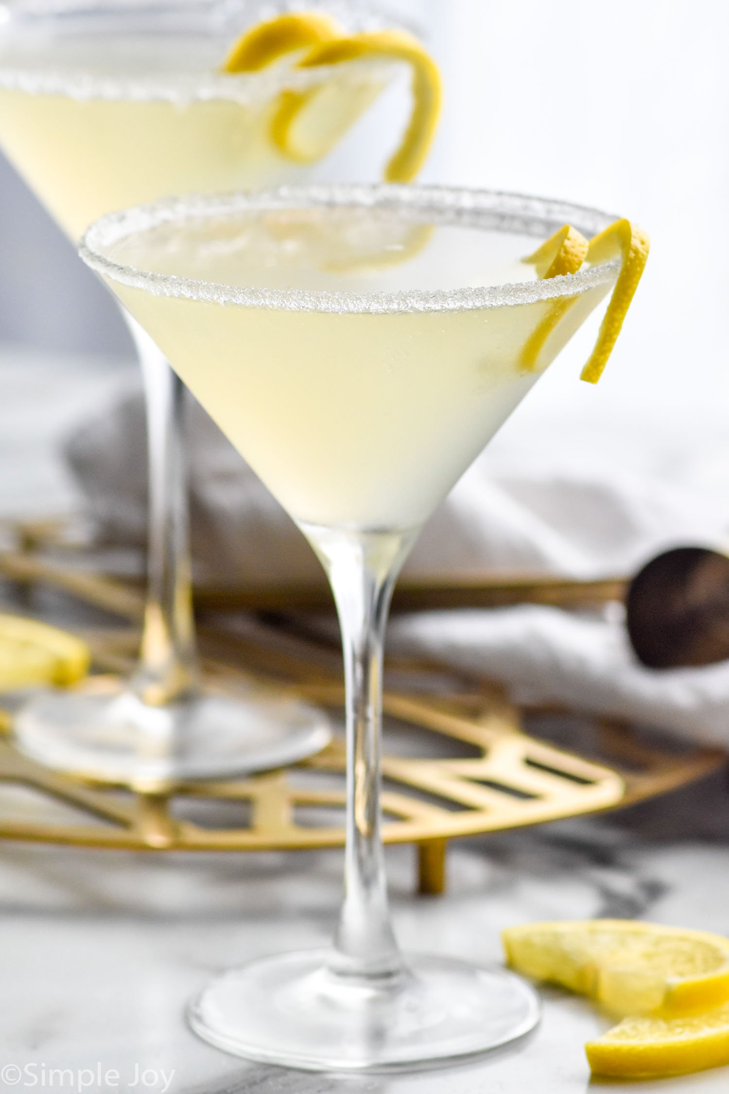 Side view of two Lemon Drop Martini cocktails garnished with lemon twist. Lemon slices beside glasses.