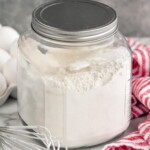 Container of homemade pancake mix. Eggs, whisk, and towel sitting beside. - 9