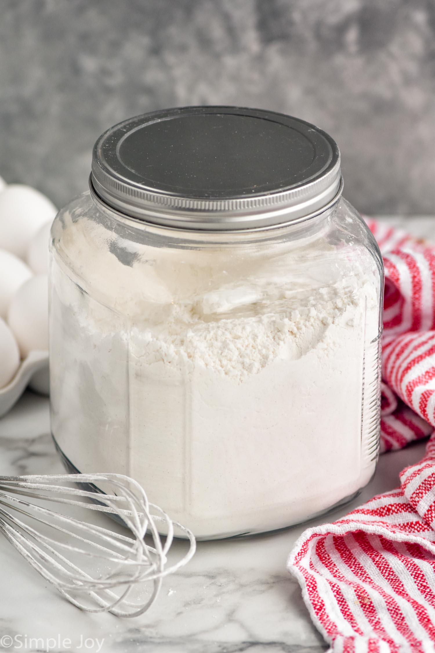 Container of homemade pancake mix. Eggs, whisk, and towel sitting beside.