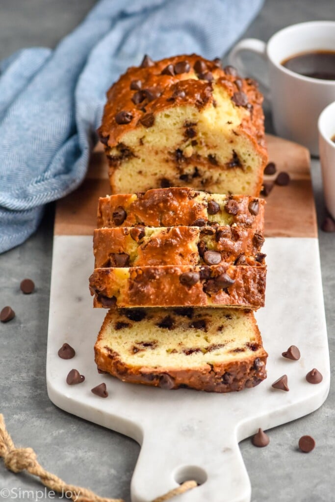 Photo of a partially sliced loaf of Chocolate Chip Bread on a cutting board with cups of coffee and chocolate chips beside.