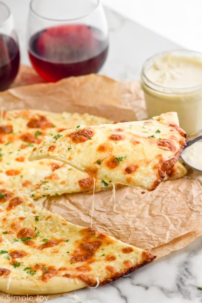 Slice of pizza with White Pizza Sauce pulling out of pizza. Jar of White Pizza Sauce and two glasses of red wine sitting beside