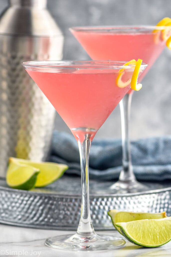 Side view of Cosmopolitan Cocktail with another Cosmopolitan Cocktail and shaker bottle on tray behind. Lime wedges beside.