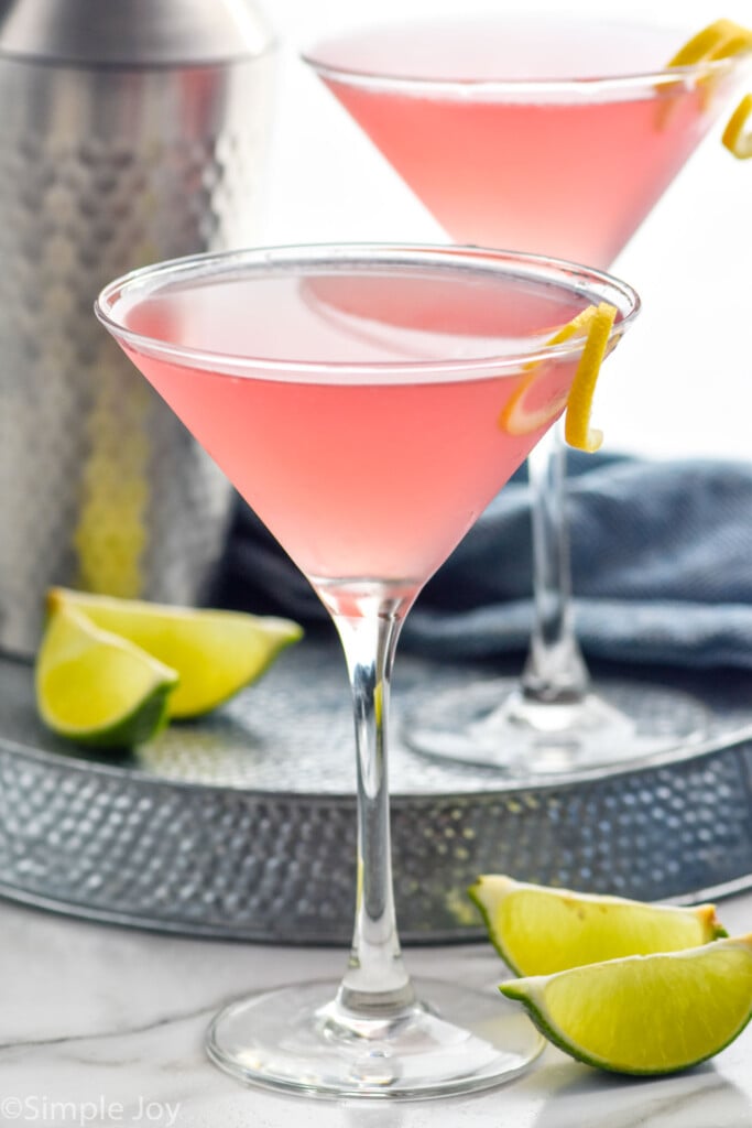 Photo of Cosmopolitan Cocktail with another cosmo and cocktail shaker behind. Lime wedges beside.