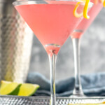 Side view of Cosmopolitan Cocktail with another cosmo and shaker bottle on tray behind. Lime wedges beside. - 8