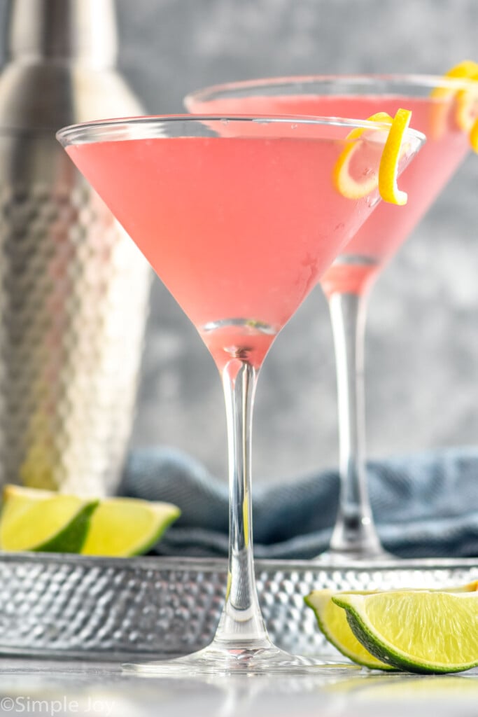 Side view of Cosmopolitan Cocktail with another cosmo and shaker bottle on tray behind. Lime wedges beside.