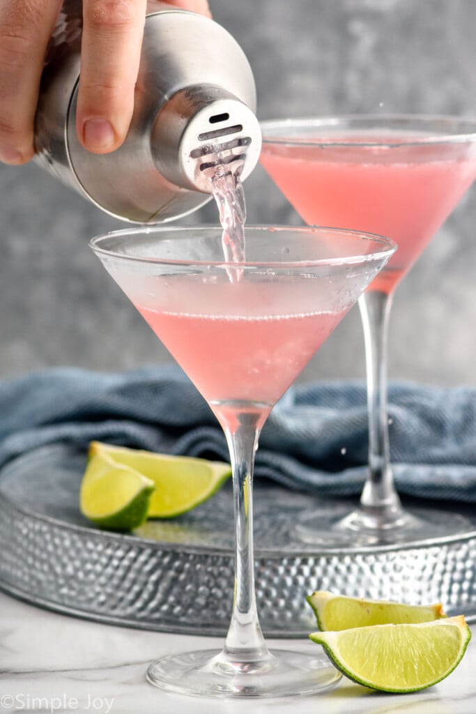 Side view of person's hand pouring cocktail shaker of Cosmopolitan Cocktail recipe into martini glasses. Lime slices beside.