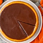 Overhead photo of a Pumpkin Cheesecake with a slice cut into it. Serving spatula, cinnamon sticks, and pumpkins beside.