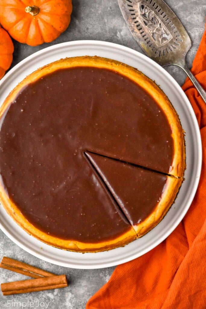Overhead photo of a Pumpkin Cheesecake with a slice cut into it. Serving spatula, cinnamon sticks, and pumpkins beside.