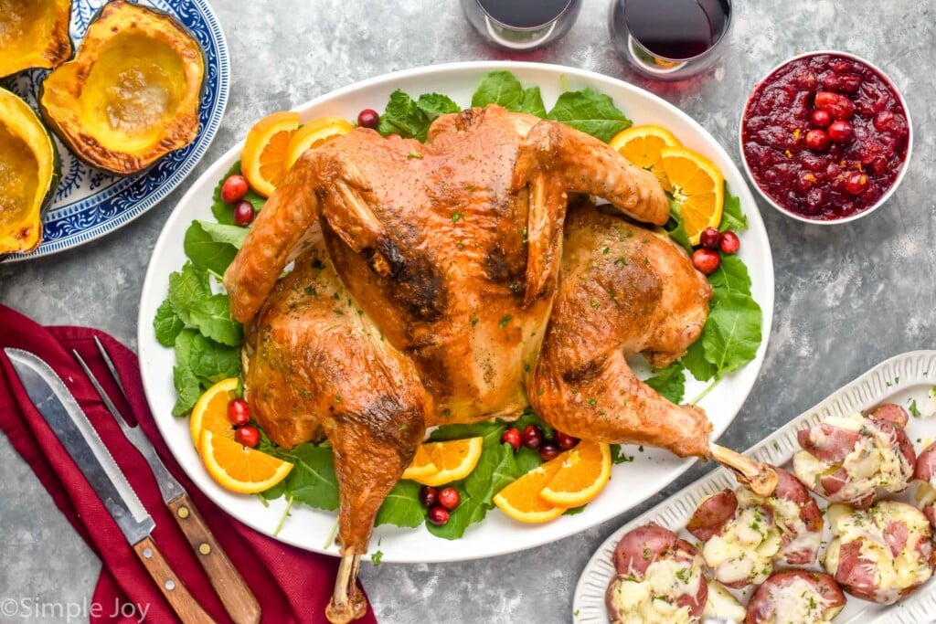 Overhead photo of Spatchcock Turkey served on a platter with greens, cranberries, and orange slices with wine, cranberry sauce, potatoes, and carving knives beside.
