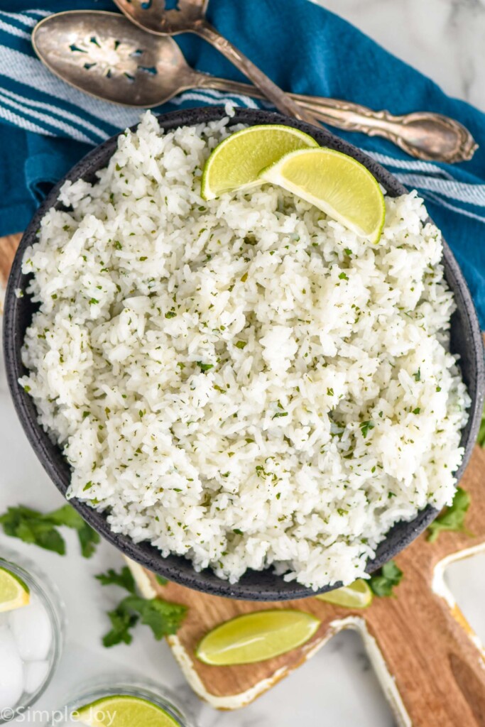 Overhead photo of a bowl of Cilantro Lime Rice with spoons, cilantro, and lime wedges beside.