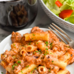 Pinterest graphic for Baked Ziti recipe. Text says, "the best Baked Ziti simplejoy.com." Image shows a plate of Baked Ziti with wine and salad beside.