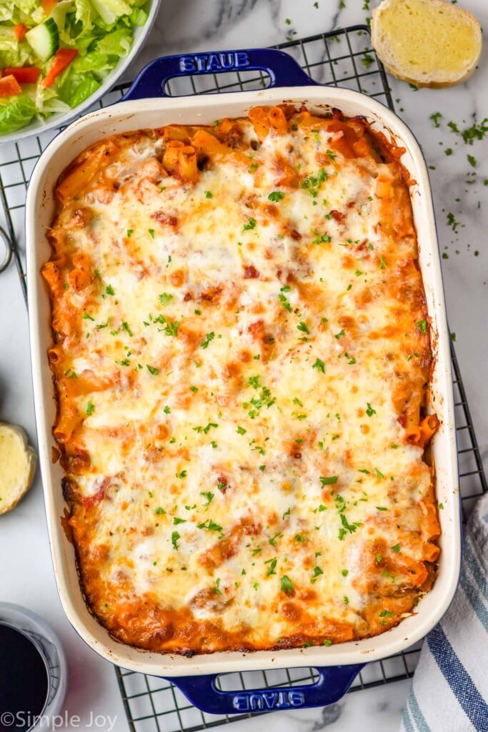 Overhead photo of a baking dish of Baked Ziti on a cooling rack. Salad and garlic bread beside.