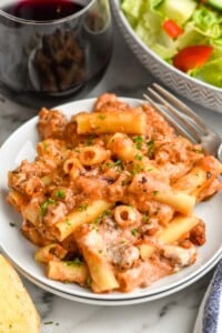 Photo of Baked Ziti served on a plate with a fork. Wine and salad beside.