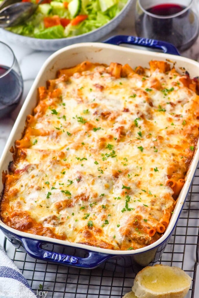 Photo of a baking dish of Baked Ziti on a cooling rack. Glasses of wine, side salad, and bread beside.