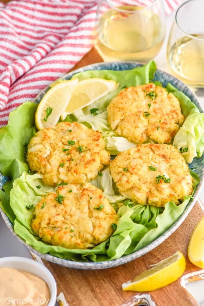 Crab Cakes on a plate with lettuce and lemon wedges. Glasses of wine and dipping sauce beside plate.
