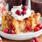 Cranberry French Toast Casserole on a plate with whipped cream and cranberries. Cups of coffee beside.