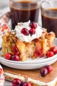 Cranberry French Toast Casserole on a plate with whipped cream and cranberries. Cups of coffee beside.