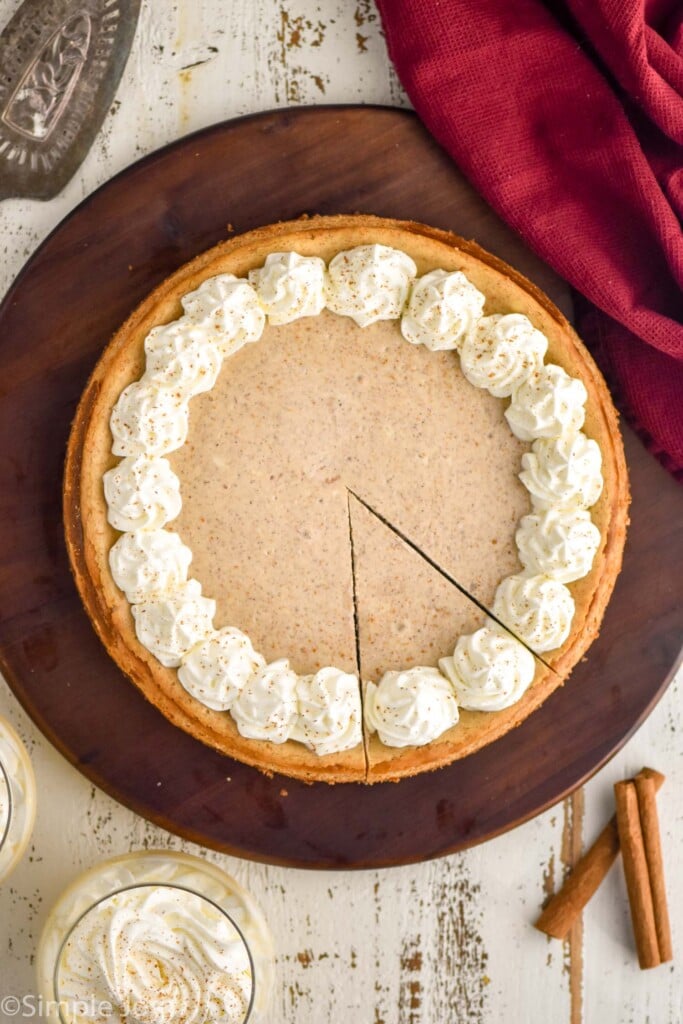 Overhead photo of Eggnog Cheesecake with one slice cut. Glass of eggnog and cinnamon sticks beside.