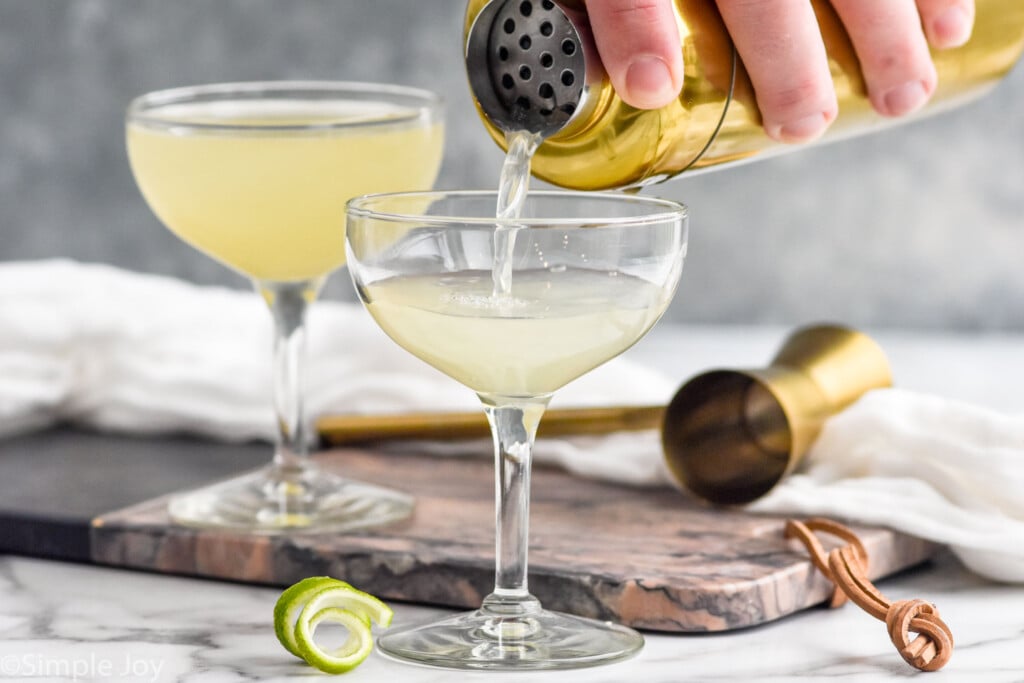 man's hand pouring a cocktail shaker of daiquiri ingredients into a glass. Glass of lime daiquiri and cocktail jigger sitting in background