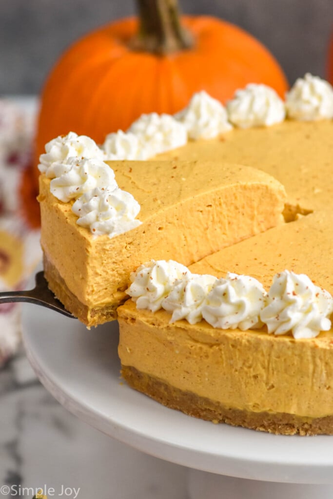 Photo of a slice of No Bake Pumpkin Cheesecake being served from No Bake Pumpkin Cheesecake on a cake plate. Pumpkin beside.