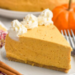 Photo of a slice of No Bake Pumpkin Cheesecake on a plate with pumpkin and cinnamon sticks beside. Whole No Bake Pumpkin Cheesecake behind. - 13