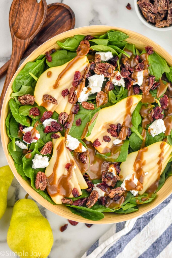 Overhead view of a bowl of Pear Salad with salad tongs, pears, and candied nuts beside.