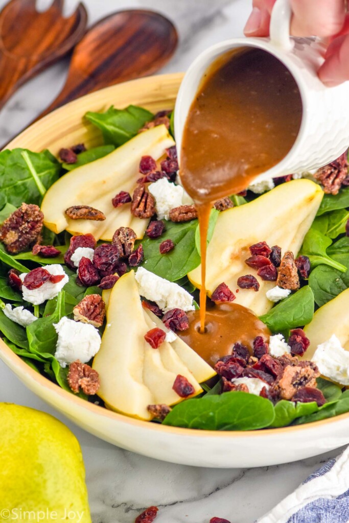 Photo of person's hand pouring dressing onto Pear Salad. Pears and salad tongs beside.