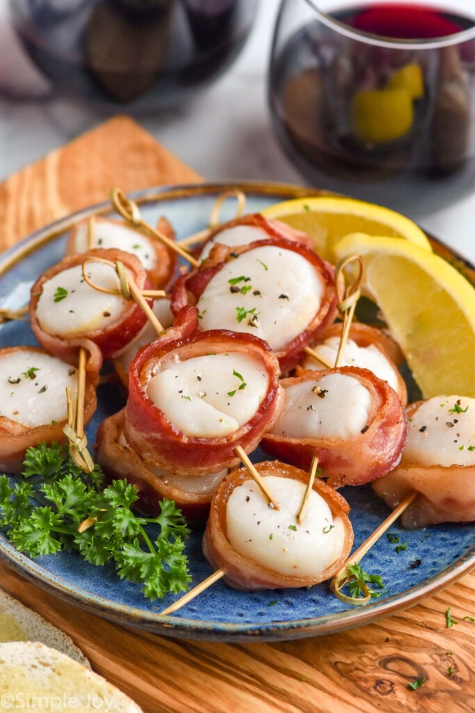 Bacon Wrapped Scallops on a plate with lemon wedges. Glasses of wine beside