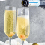 Pinterest graphic for Champagne Cocktail recipe. Text says, "the best Champagne Cocktail simplejoy.com." Image is side view of champagne being poured into champagne flutes for Champagne Cocktail recipe. Cork and cocktail jigger beside.