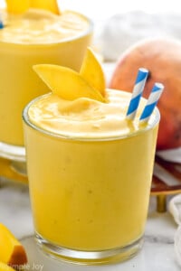 Mango Smoothie with straws and mango slice. Another Mango Smoothie and a mango beside.