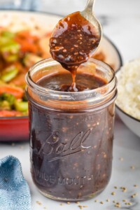 Photo of spoonful of Stir Fry Sauce over jar of Stir Fry Sauce with stir fry in skillet and bowl of rice in the background.