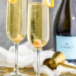 Side view of Champagne Cocktail served in champagne flutes garnished with lemon twists. Bottle of champagne, cork, cocktail jigger, and lemon twists beside. - 9