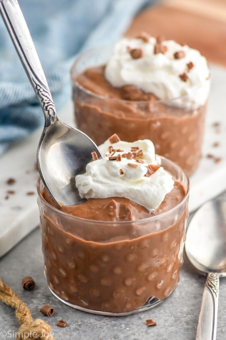 Two cups of Chocolate Pudding garnished with whipped cream and chocolate shavings. Spoon in front cup and another spoon beside