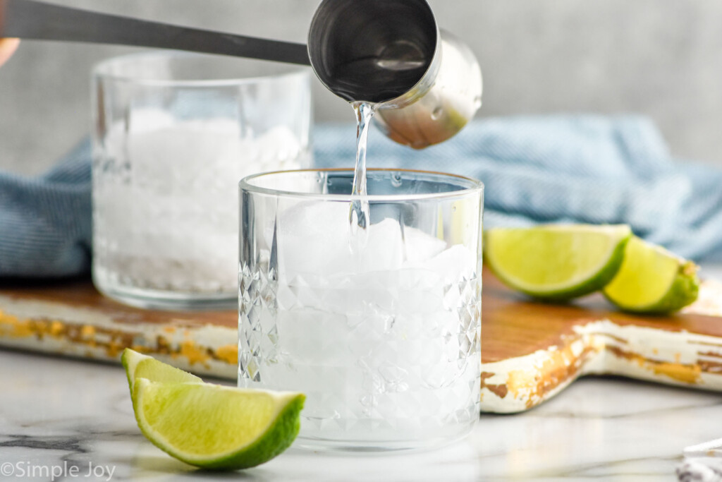 Side view of cocktail jigger of ingredient being poured into tumbler of ice for Vodka Soda recipe. Lime wedges beside.