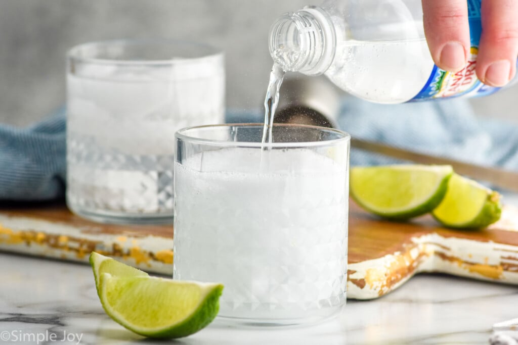 Side view of person's hand pouring club soda into tumbler for Vodka Soda recipe. Lime wedges beside.