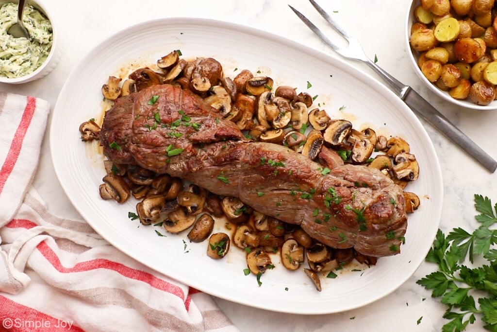 Overhead view of platter of beef tenderloin recipe serviced with mushrooms, bowl of potatoes and garlic butter beside. - 9