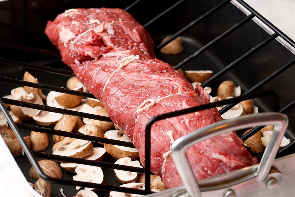 Photo of beef tenderloin salted and tied up on baking rack in pan over mushrooms before cooking. - 8