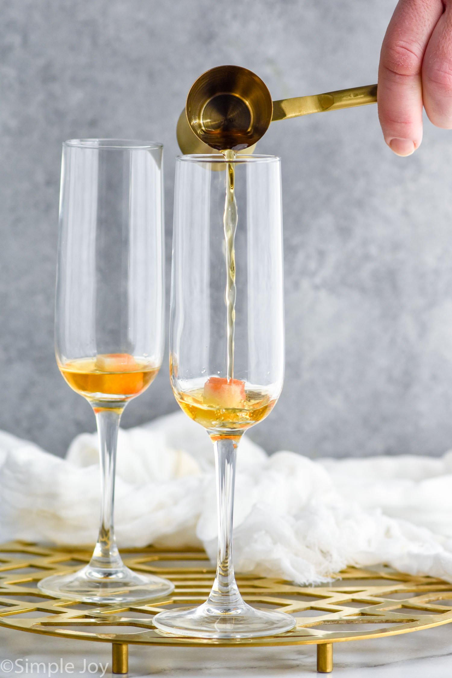 Side view of person's hand pouring cocktail jigger of ingredient into champagne flutes for Champagne Cocktail recipe.