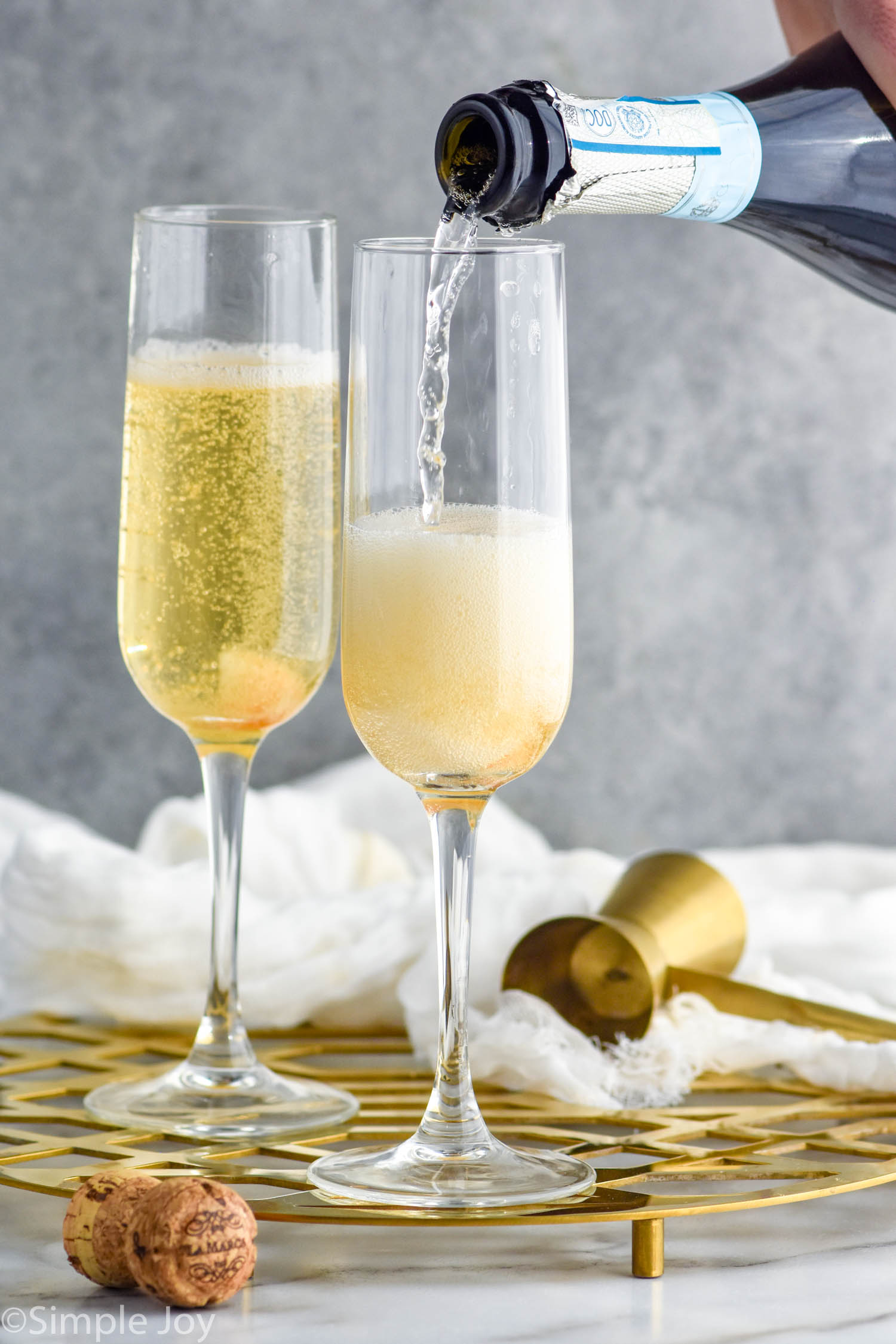 Side view of champagne being poured into champagne flutes for Champagne Cocktail recipe. Cork and cocktail jigger beside.