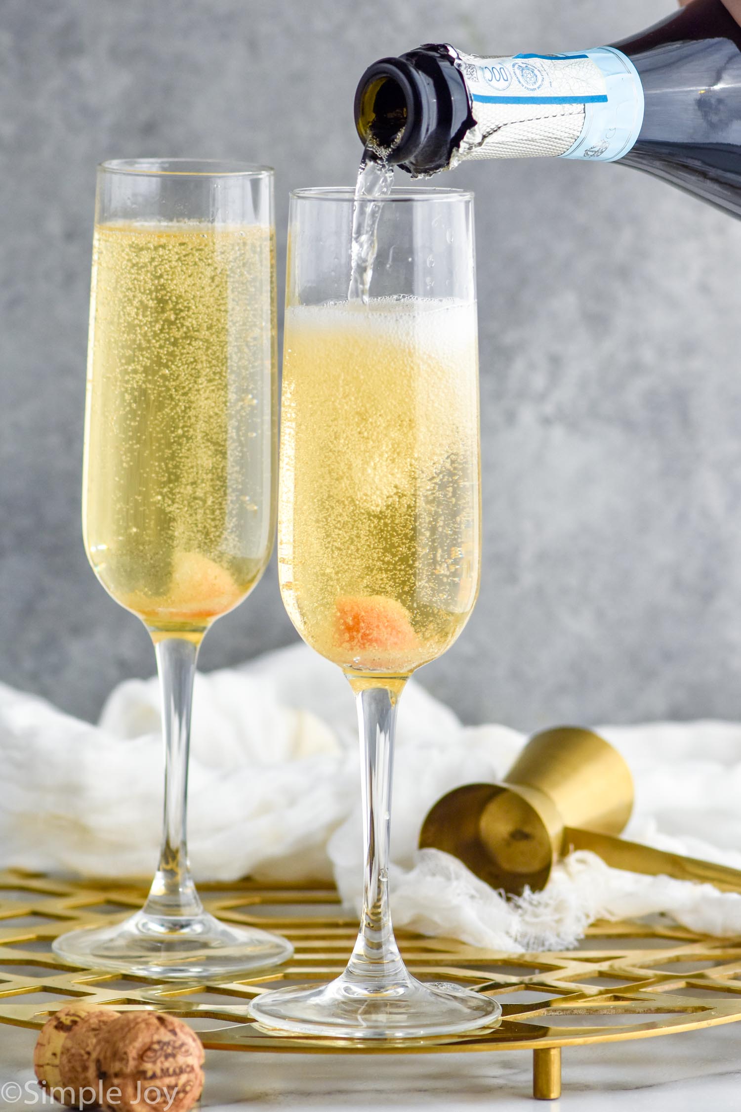 Side view of champagne being poured into champagne flutes of ingredients for Champagne Cocktail recipe. Cork and cocktail jigger beside.