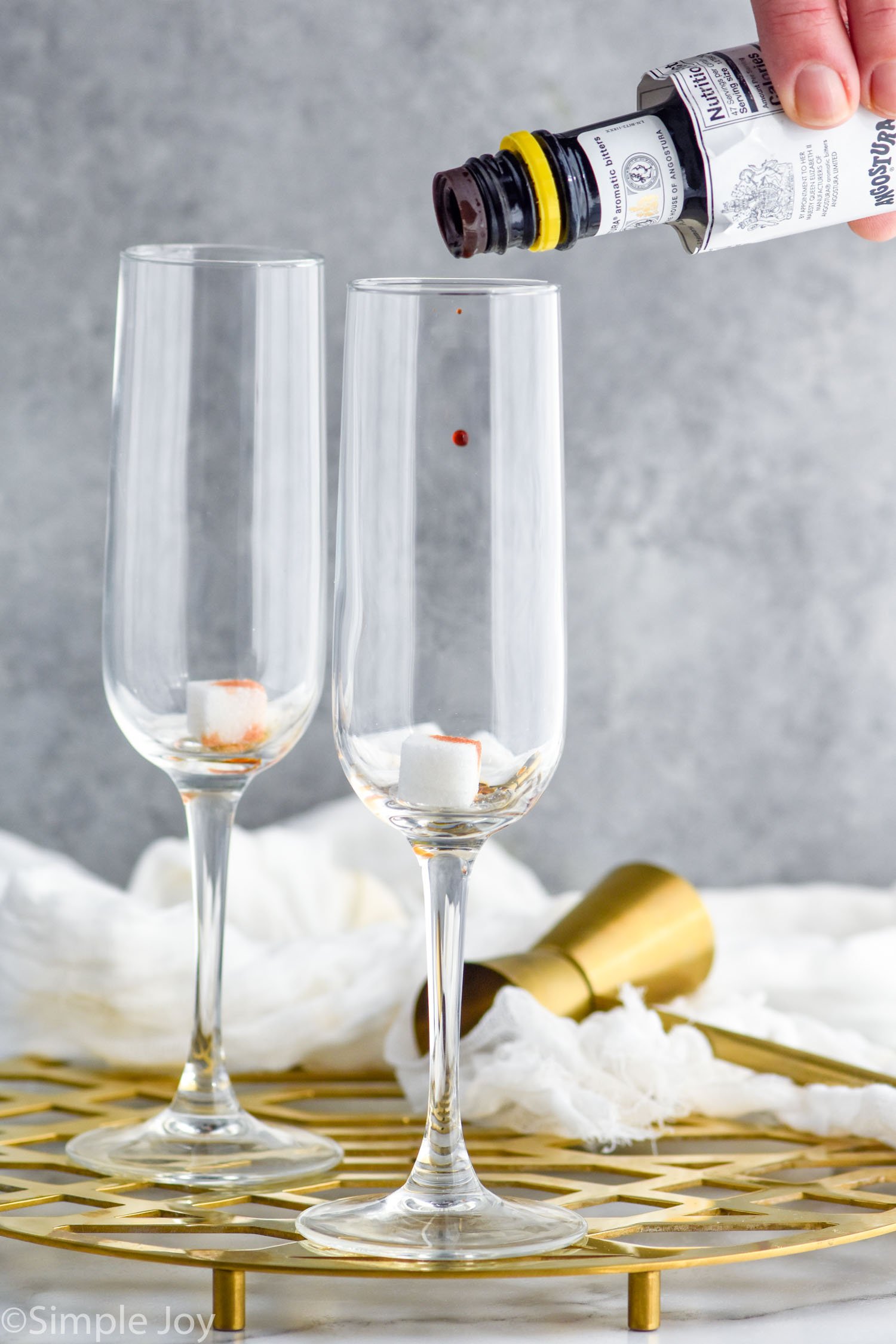 Side view of person's hand putting bitters into champagne glasses for Champagne Cocktail recipe. Cocktail jigger beside.