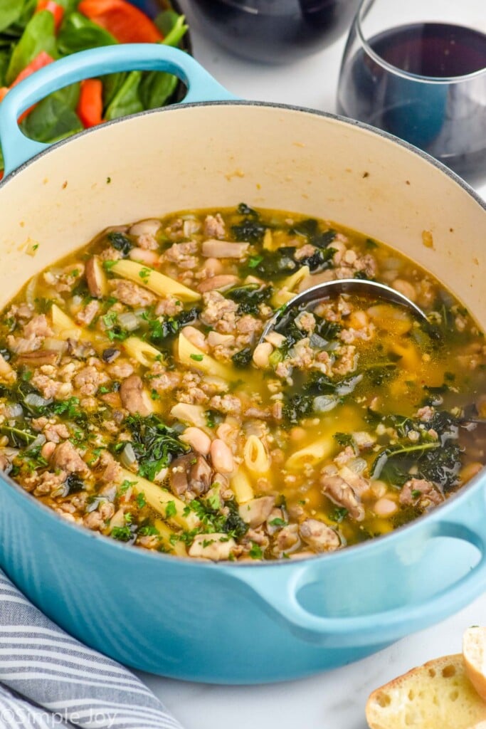 Pot of sausage and kale soup with a ladle for serving. Glasses of wine and bowl of salad beside.