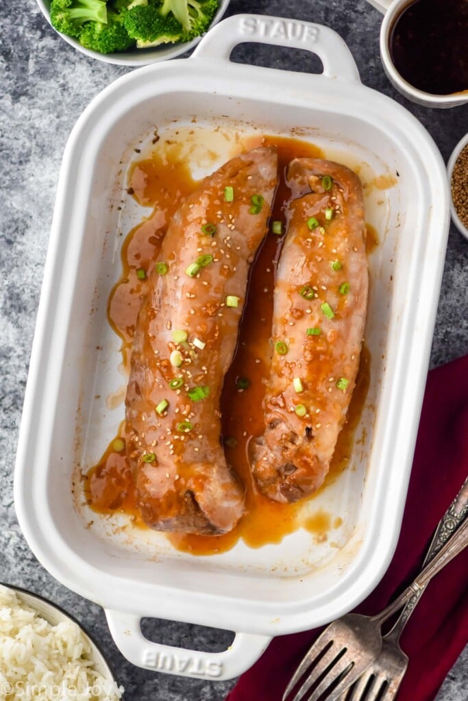 Overhead view of Teriyaki Pork Tenderloin in a baking dish. Bowl of broccoli and rice beside, as well as forks.