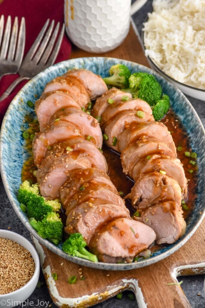 Teriyaki Pork Tenderloin sliced on a platter with broccoli. Forks, sesame seeds, and rice beside.