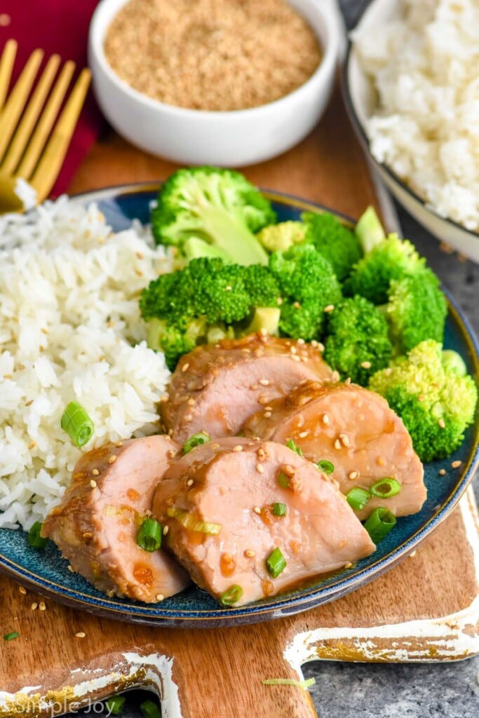 Teriyaki Pork Tenderloin on a plate with rice and broccoli. Fork and bowls of rice and sesame seeds beside.