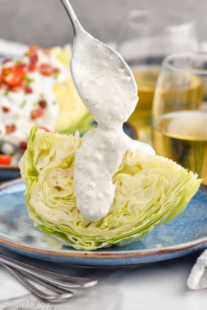 Side view of spoon of dressing over chunk of lettuce for Wedge Salad recipe. Wedge Salad and glasses of white wine beside.