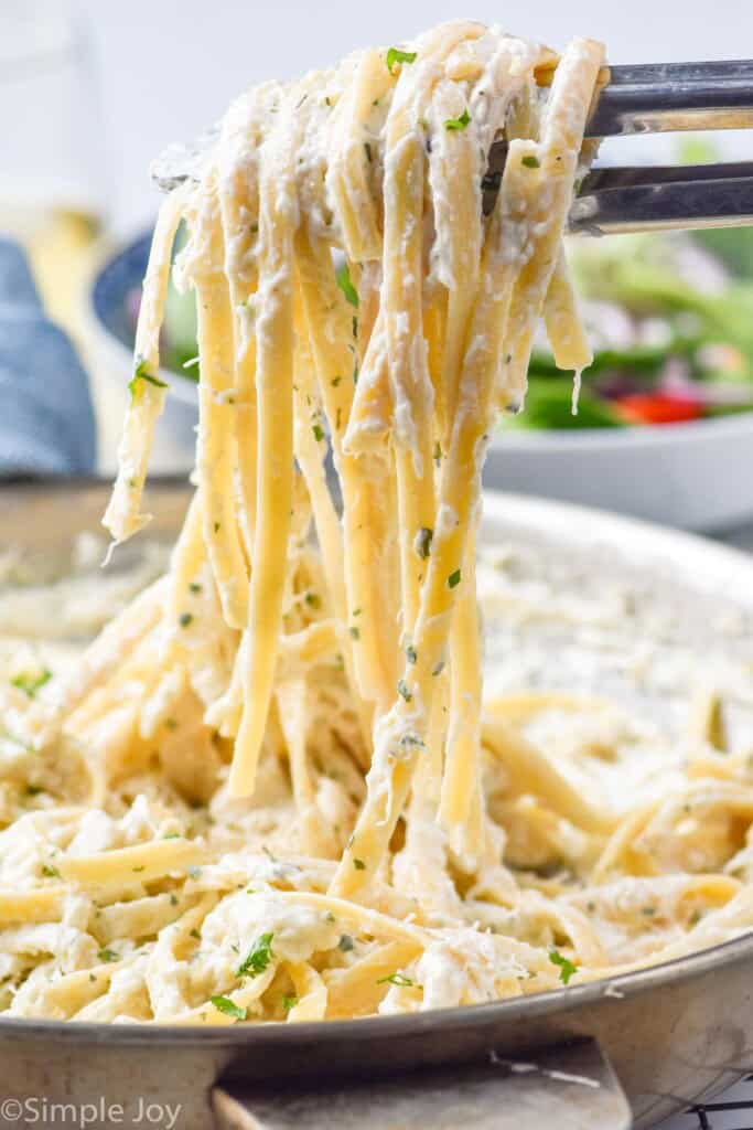 Side view of tongs lifting Creamy Crab Pasta from skillet. Bowl of salad beside.