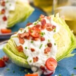 Wedge Salad served on a plate with tomatoes, bacon, and dressing. Another Wedge Salad and glasses of wine beside.