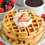 Stack of Buttermilk Waffles on a plate garnished with whipped cream and strawberry. Cups of coffee and bowl of berries beside.