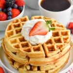 Stack of Buttermilk Waffles on a plate garnished with whipped cream and strawberry. Cups of coffee and bowl of berries beside. - 8